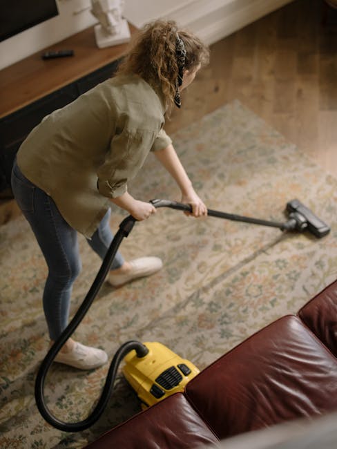 A close-up of a modern vacuum cleaner with a black floor head and a slender black handle, being used to clean a patterned, multicolored area rug with intricate floral designs. The rug is laid on a woo