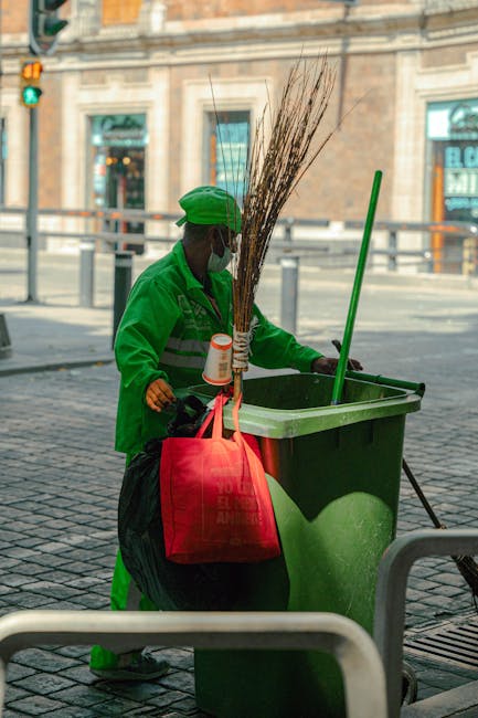 A street cleaner wearing a high-visibility orange jacket and trousers with reflective silver stripes, standing on a sidewalk during daytime in an urban environment. The worker is facing away from the