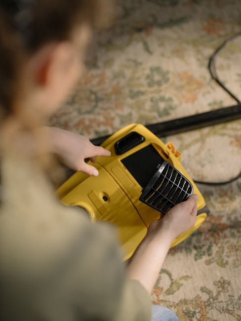 A person operating a yellow portable surface cleaning device, likely for deep carpet cleaning or sanitisation, inside a room with a patterned rug on the floor. The device features a black vent and a d