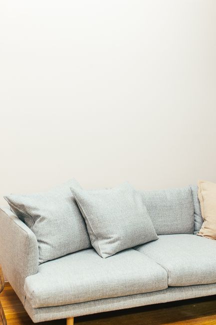 A light grey fabric sofa with multiple cushions positioned on a wooden floor against a plain white wall, featuring a beige cushion among the grey ones. The sofa appears clean and well-maintained, refl