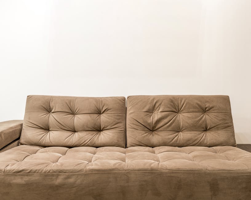 A light grey fabric sofa with multiple cushions positioned on a wooden floor against a plain white wall, featuring a beige cushion among the grey ones. The sofa appears clean and well-maintained, refl